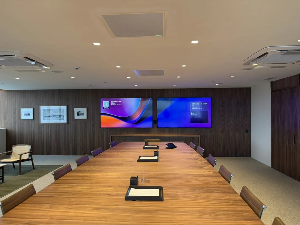 Fotografia da sala de reunião usada como referência, com mesa de madeira e duas telas na parede frontal.